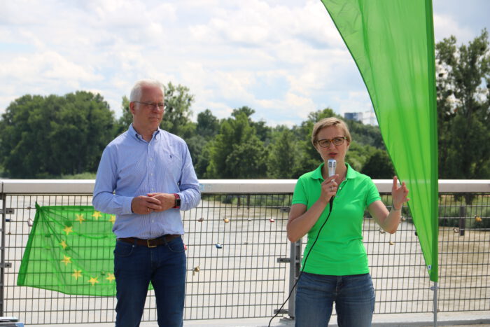 Franziska Brantner und Thomas Zawalski stehen vor Fahnen der Grünen auf der Passerelle zwischen Kehl und Straßburg.